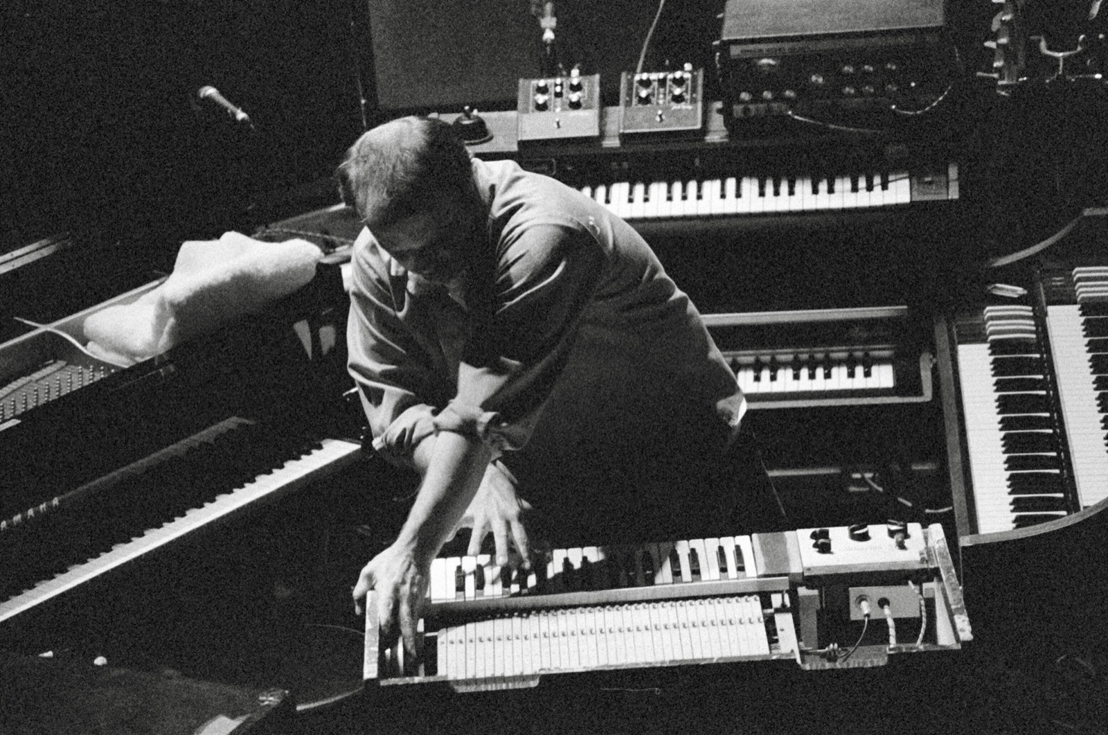 A keyboardist hunches low over a clavinet in the middle of a ring of keyboards, shot from above in black and white.